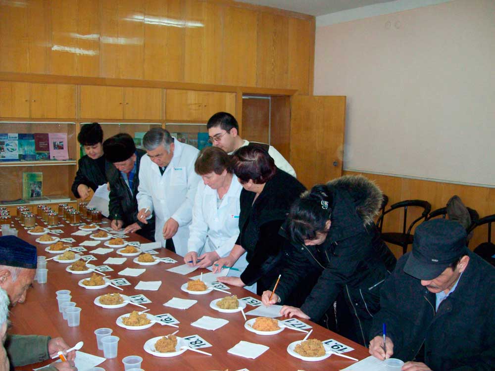 Ein farbiges Foto, das drei Labormitarbeiter in weißen Kitteln und sechs Probanden in dunkler Alltagskleidung beim Testen von Speisen zeigt.