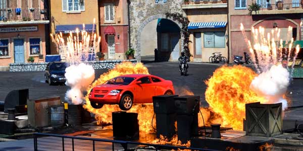 Farbiges Szenenfoto eines Actionsfilms für die-besten-spielfilme: Bei einer Explosion auf einem städtischen Platz fliegt ein roter Sportwagen in die Luft.
