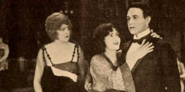 Schwarzweiß Szenenfoto eines alten Hollywoodfilms für die-besten-spielfilme: Unter den finsteren Blicken einer Nebenbuhlerin schmiegt sich eine strahlende Frau an die Schulter eines ernst dreinblickenden Mannes.